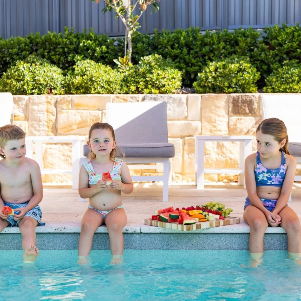 Three children sitting on the edge of a pool with a fruit platter behind them